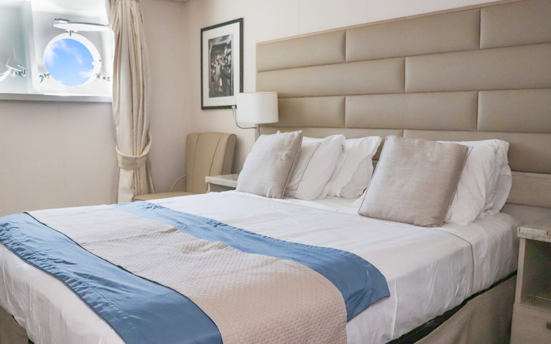 Porthole Stateroom with two portholes, vanity, stool, nightstand, chairs & small table next to bed fitted in white & tan linens.
