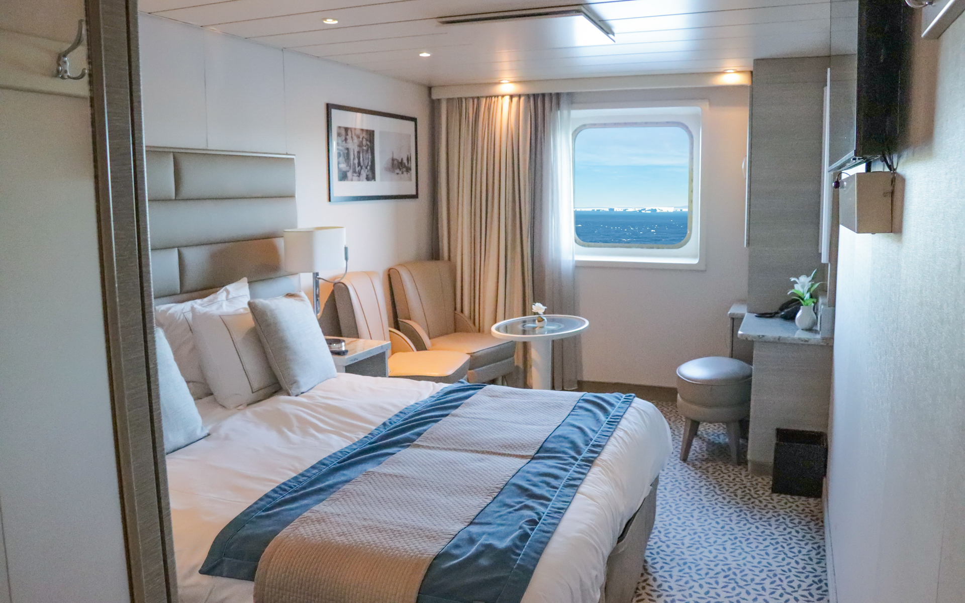 Solo Vista Stateroom cabin aboard Discoverer: Polar Latitudes with large viewing window, chairs & small table next to a bed fitted in white and tan linens.