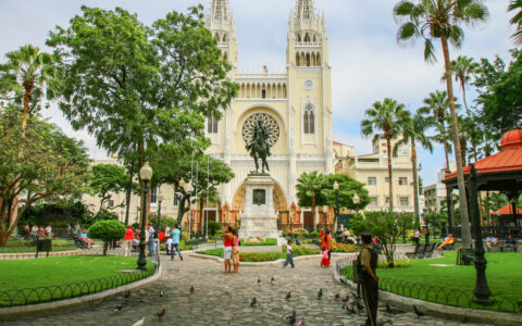 Yellow church with large circular stained glass window beside grassy park with cobblestone walkways & Ecuador travelers.