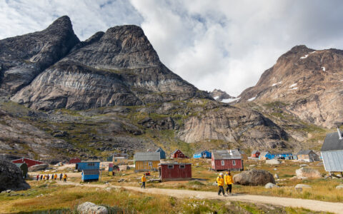 Small group of Greenland Explorer cruise travelers walk a dirt road beside colorful & weathered homes in a remote village.