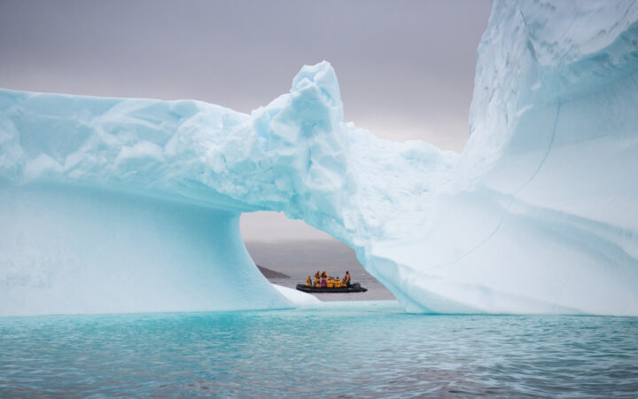 Zodiac with Greenland Explorer cruise travelers peeks through a hole in an a large blue iceberg in calm sea under cloudy skies.