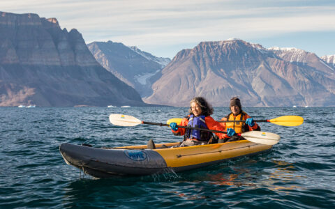 Tandem kayakers paddle a yellow inflatable boat in calm water with brown mountains behind on the Greenland Explorer cruise.