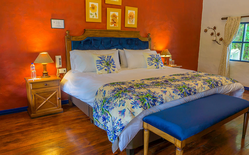 Wooden double bed with bench in floral sheets at Hacienda Pinsaqui with wood floors, window & red & white walls.