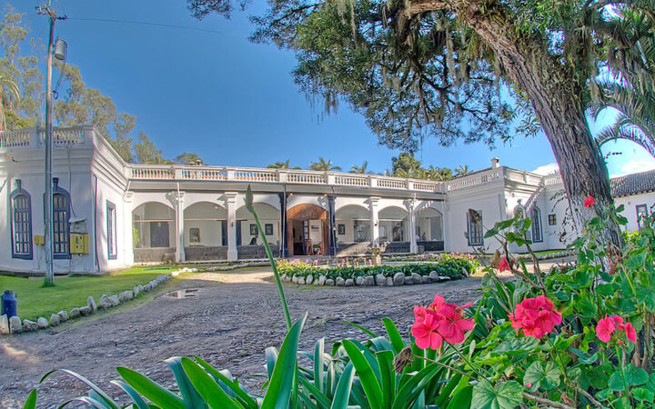 Exterior of colonial white 1-story building at Hacienda Pinsaqui by cobblestone rotunda & bright gardens under a blue sky.