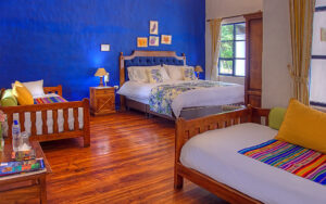 Wooden double bed in floral sheets & 2 wooden daybeds at Hacienda Pinsaqui with wood floors, window & blue & white walls.
