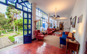 Wall of windows opening onto garden courtyard at Hacienda Pinsaqui, with rosy stone tiles, red & blue upholstered chairs & art.