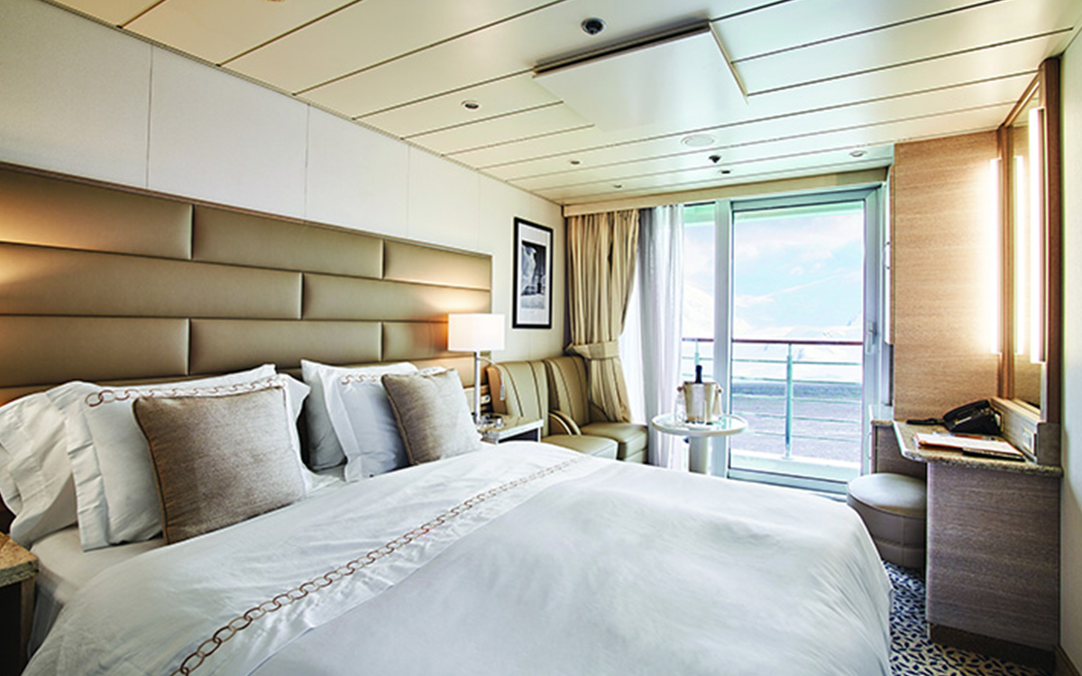 Balcony Stateroom on small ship showing a large bed fitted with white & tan linens next to chairs, desk & sliding glass balcony doors