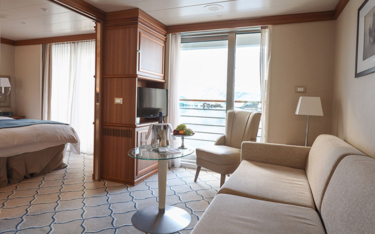 Spacious Discovery Suite aboard small ship with bed to the left & couch with chair & table to the right separated by TV cabinetry