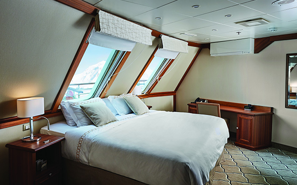 Grand Suite complimented by wood accents and two overhanging windows shows bed fitted with white & tan linens aboard small ship.