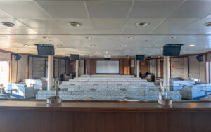 Spacious lecture room aboard small ship with several rows of couches facing front of room with projector screen at the center