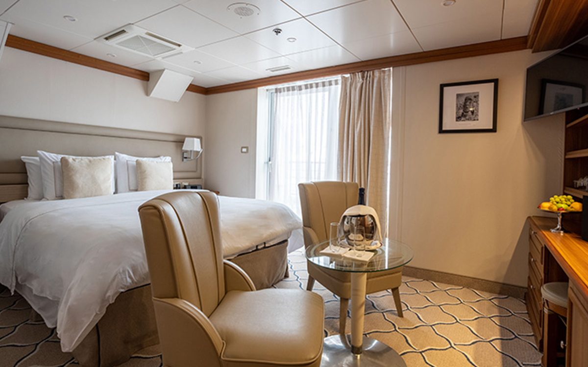 Navigator Stateroom boasts large bed fitted with white & tan linens next to large tall window and seating at its foot aboard small ship.