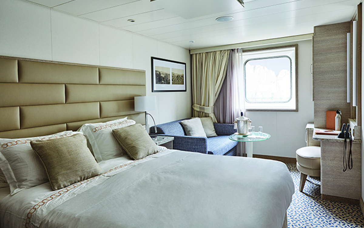 Ocean Stateroom on small ship showing a large bed fitted with white & tan linens next to couch, desk, vanity, stool & large window