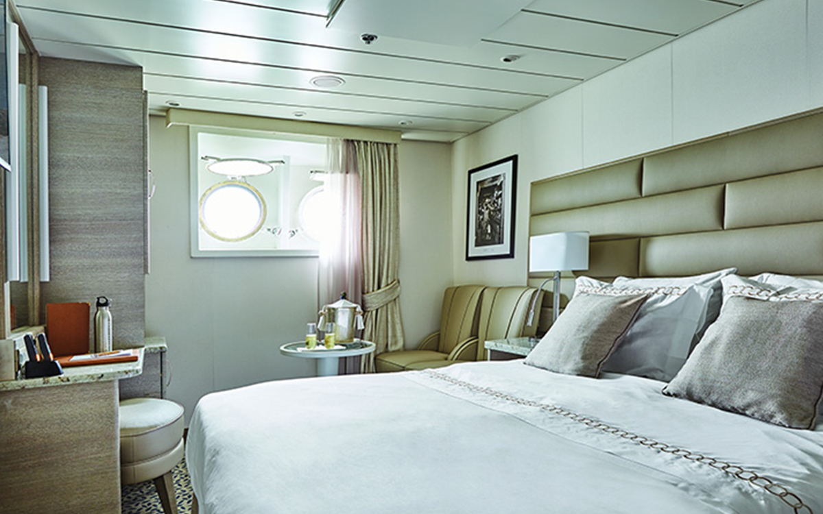 Porthole Stateroom with two portholes, vanity, stool, nightstand, chairs & small table next to bed fitted in white & tan linens.