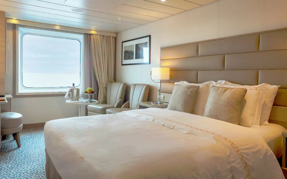 Solo Vista Stateroom cabin with large viewing window, chairs & small table next to a bed fitted in white and tan linens.