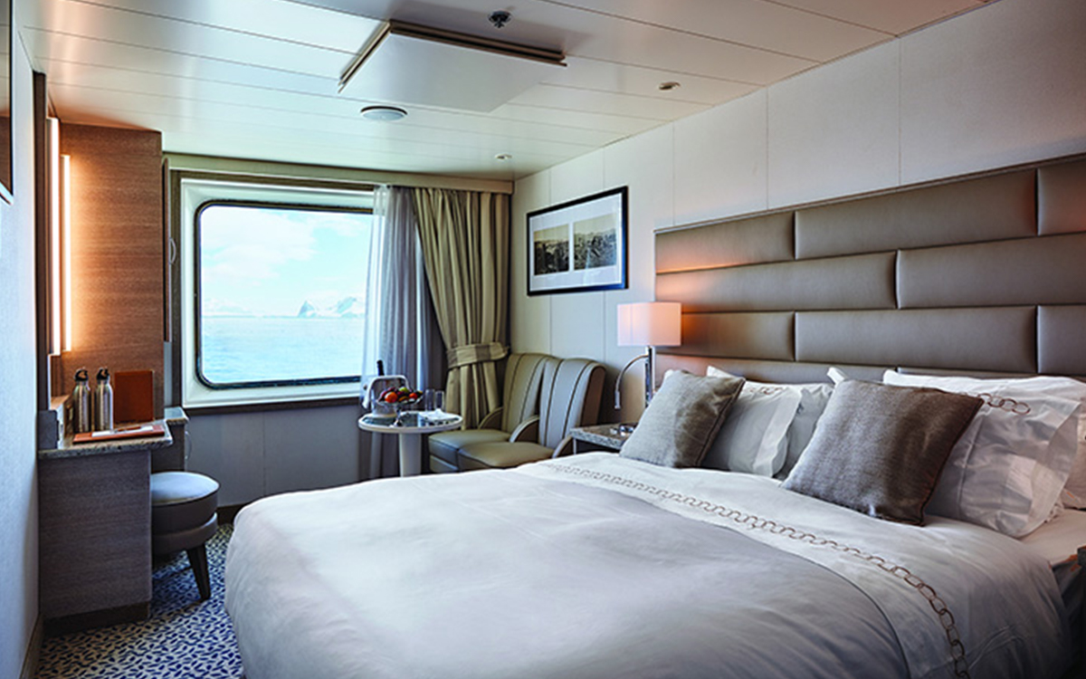 Vista Stateroom with large viewing window, vanity, stool, chairs & small table next to a bed fitted in white and tan linens.