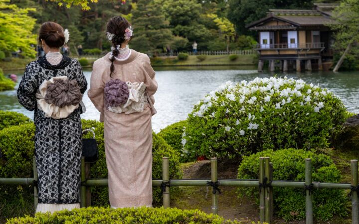 2 Japanese women in traditional garb tied in large bows on their backs stand among lush green gardens overlooking a pond.