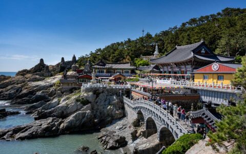Seaside South Korean town with colorful buildings backed by green forest overlooking boulders leading into the sea in the sun.