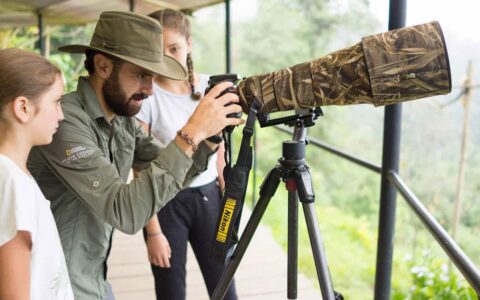 Man in green button-down shirt & hat adjusts camera with long camouflage lens sitting atop a tripod, as 2 girls look on.