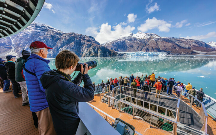 Cruise guests gather at the ships bow to take photos of a large glacier that extends to the water from an Alaska mountain range.