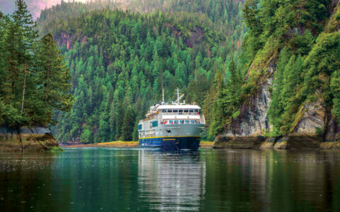 Nat Geo Quest small ship with blue hull & white upper decks cruises on glassy water between lush green mountains.