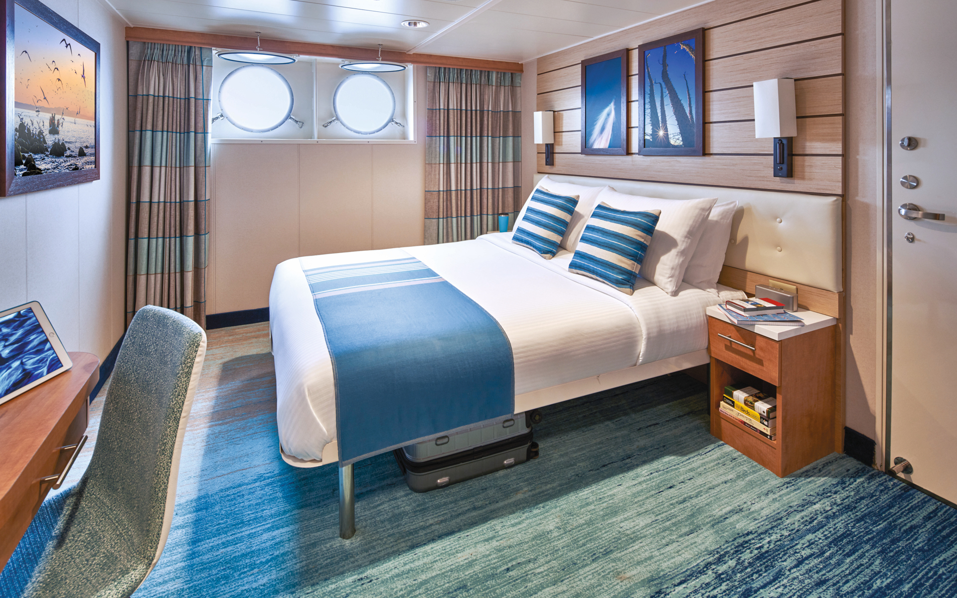 Category 2 cabin aboard Nat Geo Venture & Quest with queen bed in white sheets & duvet, 2 portholes, wooden desk & side table.