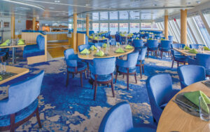 Window-lined dining room aboard National Geographic Venture with blue chairs and booths with tables set for a meal and buffet in the back.