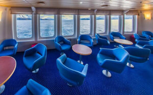 Saloon aboard small ship Ocean Nova with multiple deep blue chairs and small tabled placed throughout the room next to window-lined wall