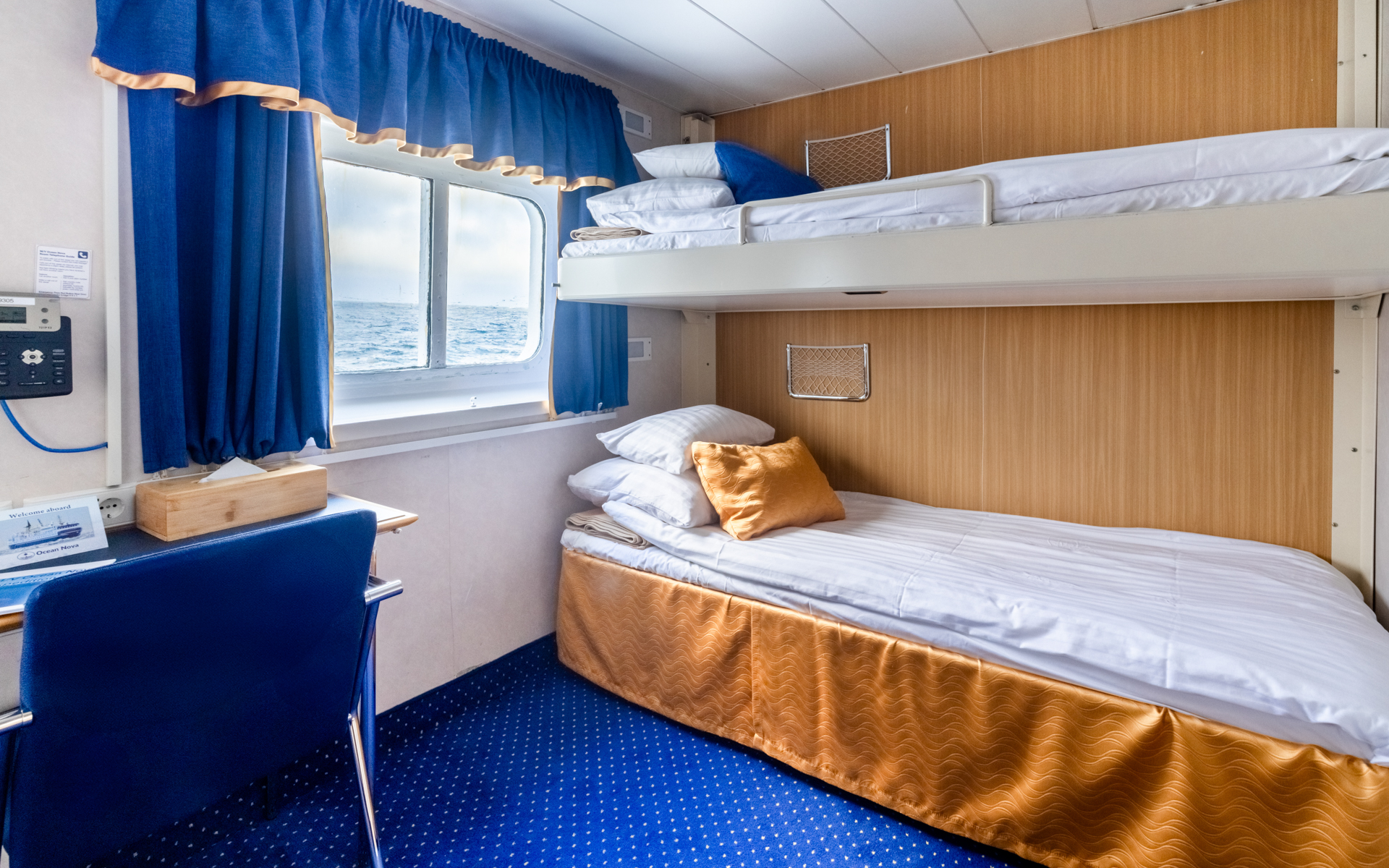 1 upper & 1 lower twin bed in white linens in classic twin cabin with blue & gold accents, small desk & armoire on Ocean Nova ship.