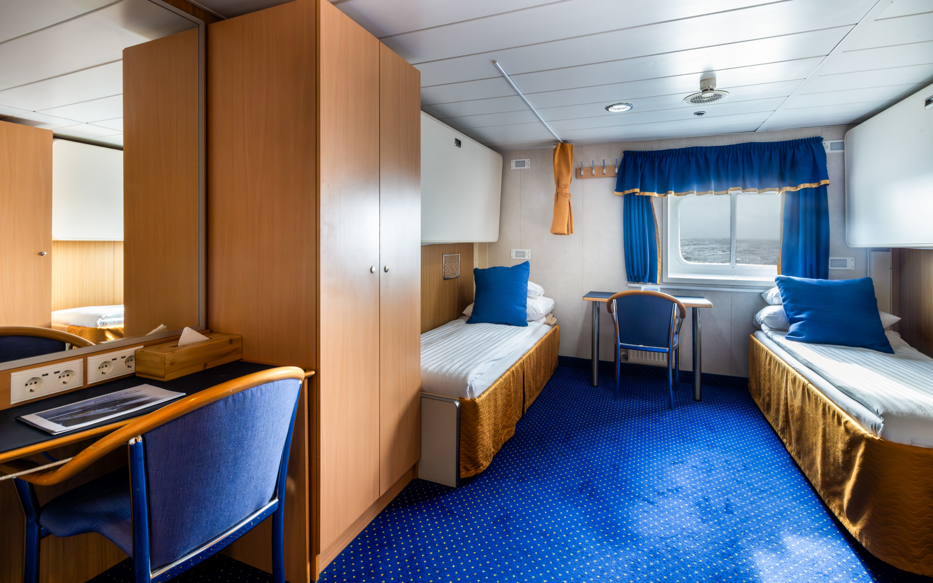 2 lower twin beds in white linens in comfort twin cabin with blue & gold accents, small desk & armoire on Ocean Nova ship.