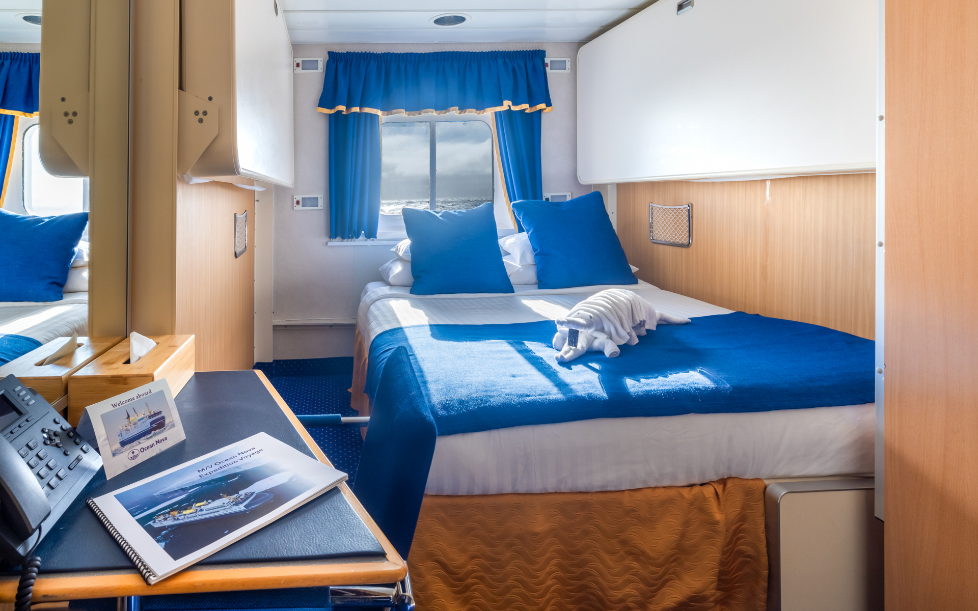 Double bed in white linens in explorer double cabin with blue & gold accents, small desk & wooden armoire on Ocean Nova ship.