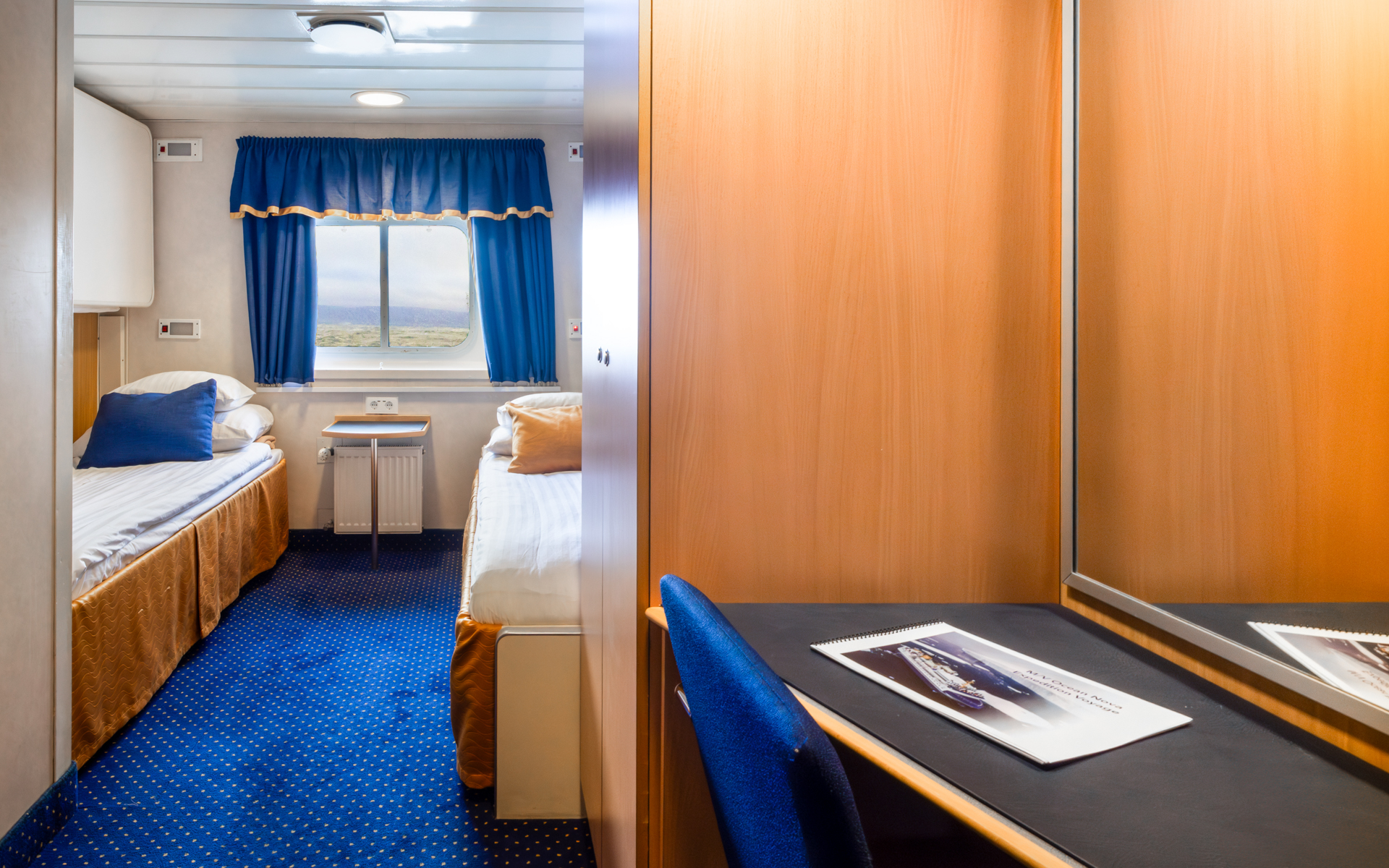 2 lower twin beds in white linens in explorer twin cabin with blue & gold accents, small desk & armoire on Ocean Nova ship.