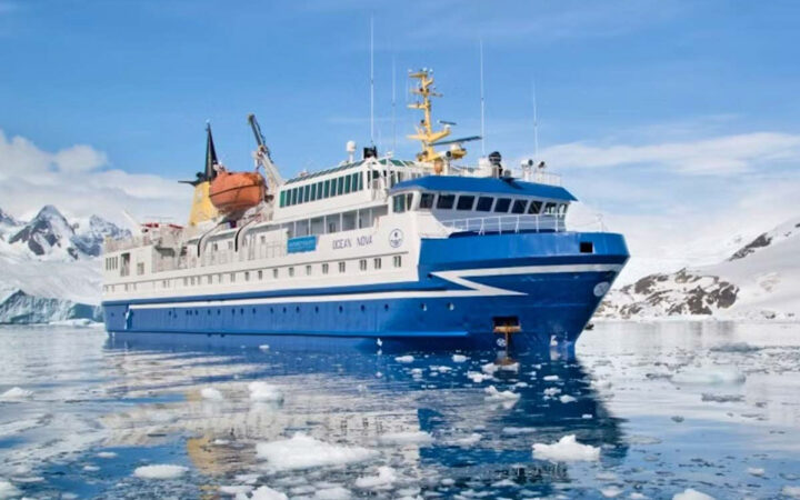Ocean Nova expedition ship with blue hull, white upper decks & orange lifeboat sits in glassy water beside snowy Antarctic cliffs.