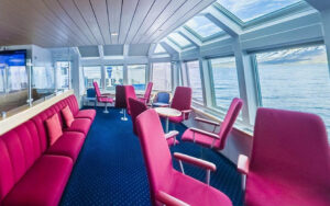 Panorama Lounge on Ocean Nova expedition ship with walls of view windows, tall red seats & benches, small tables & blue carpet.