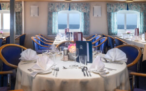 Dining room on Ocean Nova polar ship with view windows & 4- & 6-top tables set in white linens & upscale dinnerware.