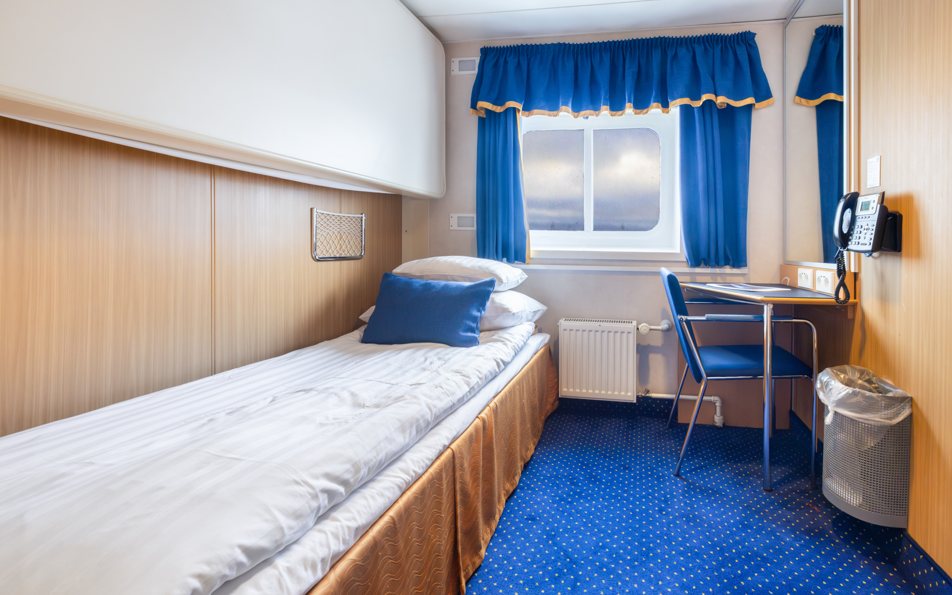 1 lower twin bed in white linens in single cabin with blue & gold accents, small desk & armoire on Ocean Nova ship.