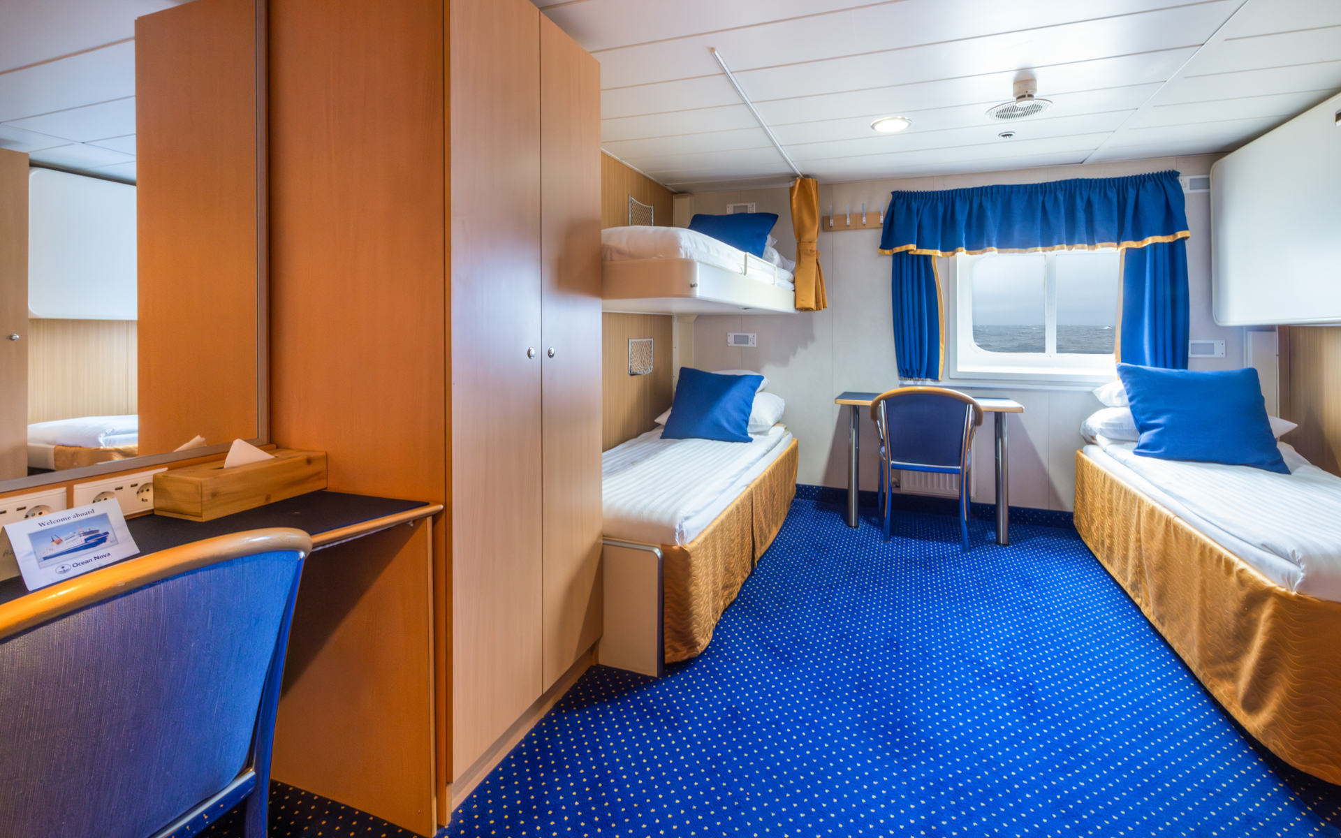 2 lower & 1 upper twin bed in white linens in triple cabin with blue & gold accents, small desk & armoire on Ocean Nova ship.