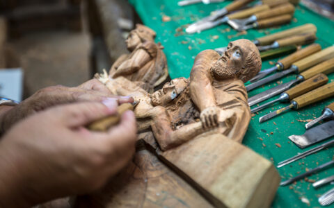 Artisan carves small wooden statues with various knives laid out on a green workbench, seen on the Otavalo Escape tour in Ecuador.