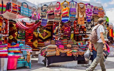 Young traveler with backpack walks past brightly colored market stall with woven goods on the Otavalo Escape tour in Ecuador.