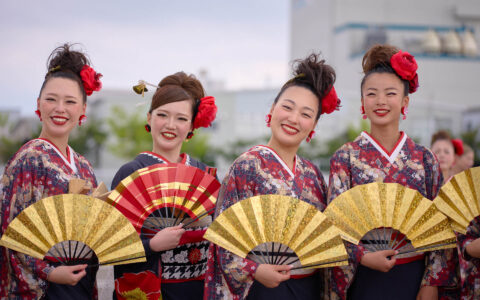 4 Japanese women in red & purple kimonos with crimson flowers in their hair & gold fans in their hands smile for a photo.