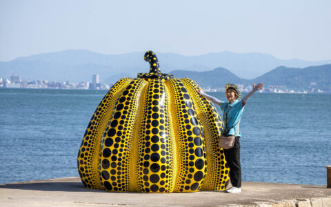 Japanese female guide in blue shirt & black pants waves beside human-size yellow & black polka dotted pumpkin by the sea.