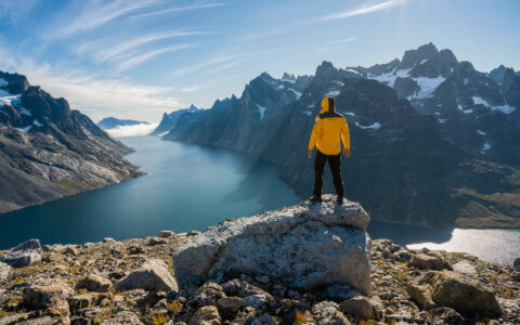 Greenland adventure cruise traveler stands atop mountain's edge looking down onto idyllic fjord among jagged mountains in sun.