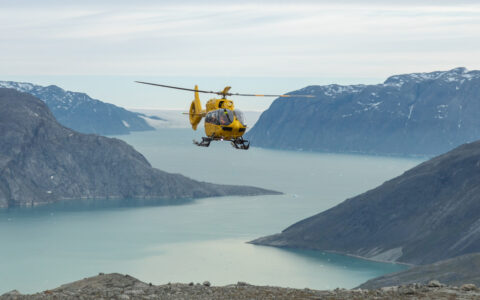 Yellow helicopter flies high above large fjord with glacier & smooth mountains during a South Greenland adventure cruise.