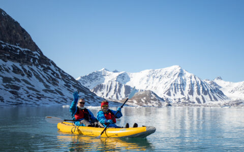 Two travelers in life jackets on a paddle excursion in calm waters in front of massive snow-covered mountains in Greenland