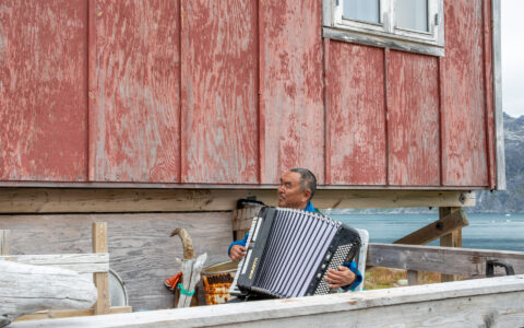 Greenland local in a blue shirt playing an accordion next to a red-painted wooden building with wood fencing in foreground.