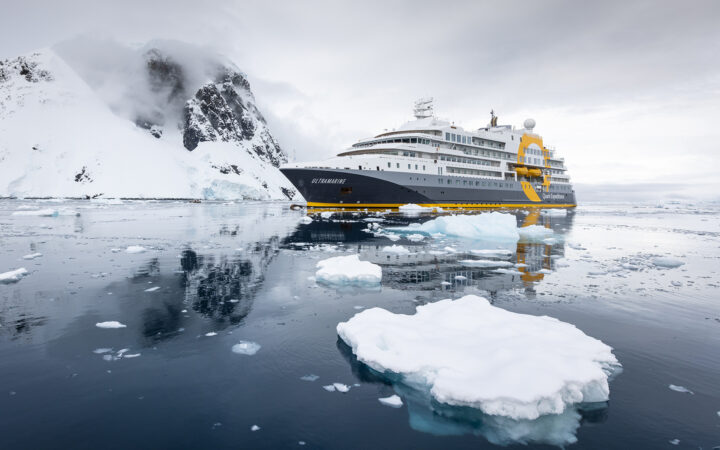 Exterior of bow of Ultramarine polar expedition ship, with snowy peaks beside & small floating icebergs in glassy, dark ocean.