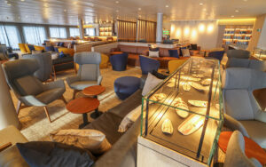 Lounge with modern blue and yellow swivel chairs, leather couches, view windows & artifacts in a glass case on Ultramarine ship.