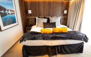 Double bed on Ultramarine ship with white sheets & duvet, gray fleece blanket, black bed skirts, wood headboard & large window.