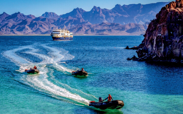 3 black inflatable Zodiac boats cruise away from Nat Geo Venture ship through turquoise waters near rocky coastline in Baja.