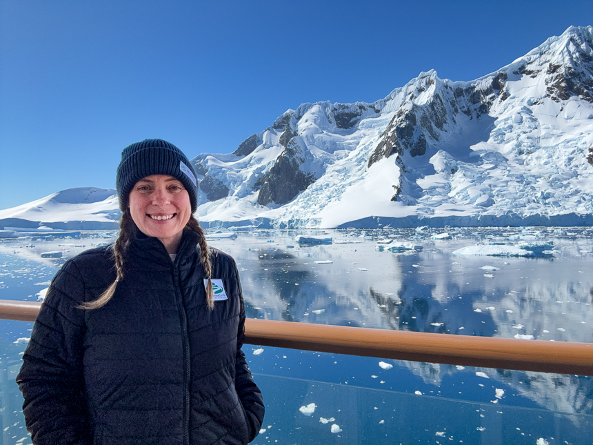 AdventureSmith explorations expert Jenn is standing aboard Sylvia Earle in front of a beautiful snowy landscape in Antarctica