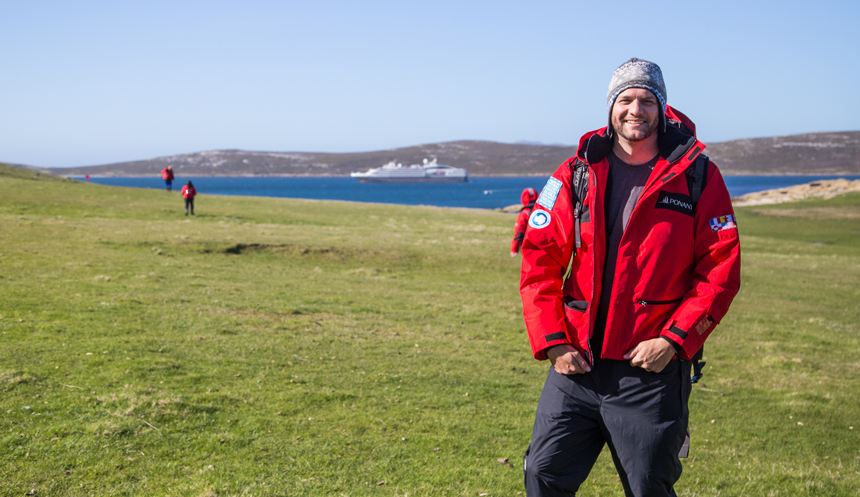 Man in red parka stands atop grassy field beside blue bay with small expedition ship waiting on a sunny day.